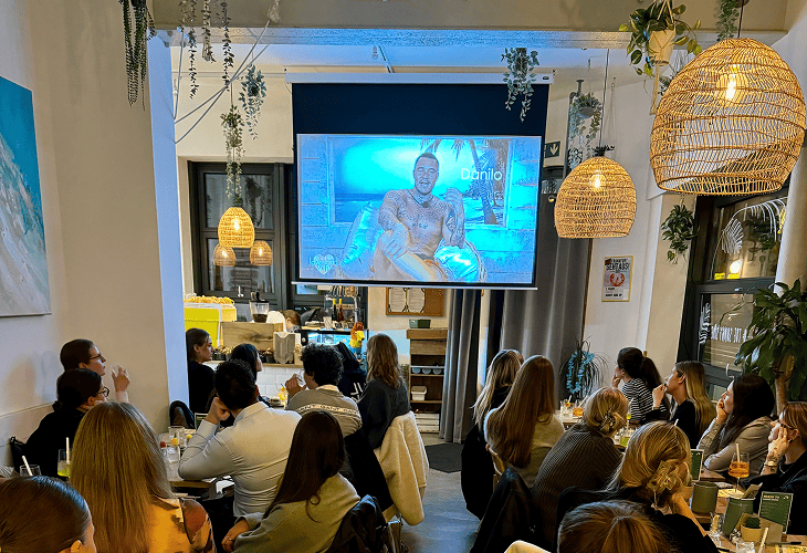 Menschen sitzen in einem Restaurant und schauen auf einen Projektor, der eine Person auf einem Bildschirm zeigt.