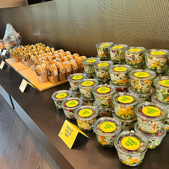 Eine Präsentation von Salatgläsern und Sandwiches auf einem Tisch. Die Gläser sind mit gelben Etiketten beschriftet.
