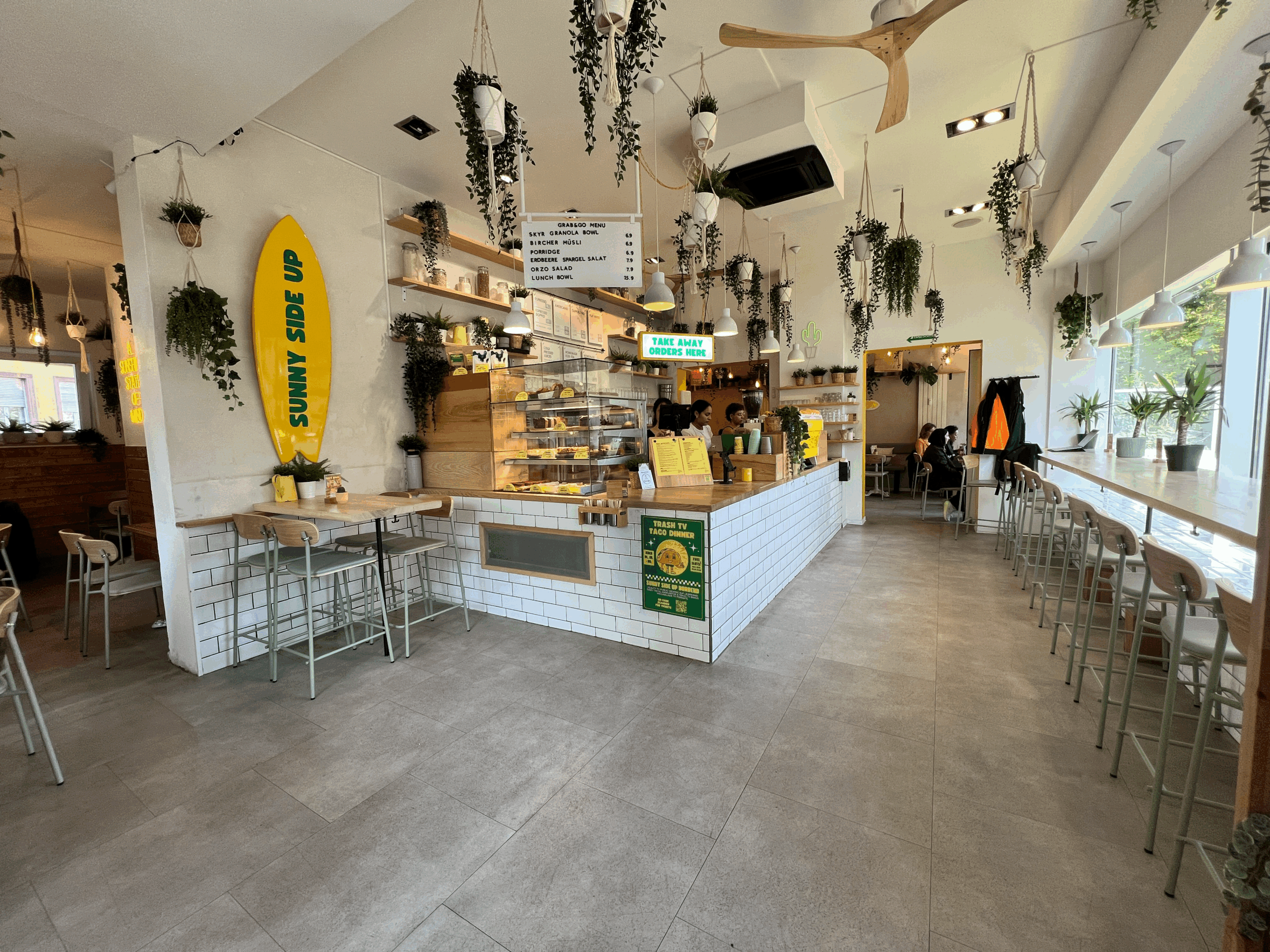 Helles Café mit minimalistischer Einrichtung, Pflanzen, einer Surfbrett-Dekoration und einer Theke für Bestellungen.
