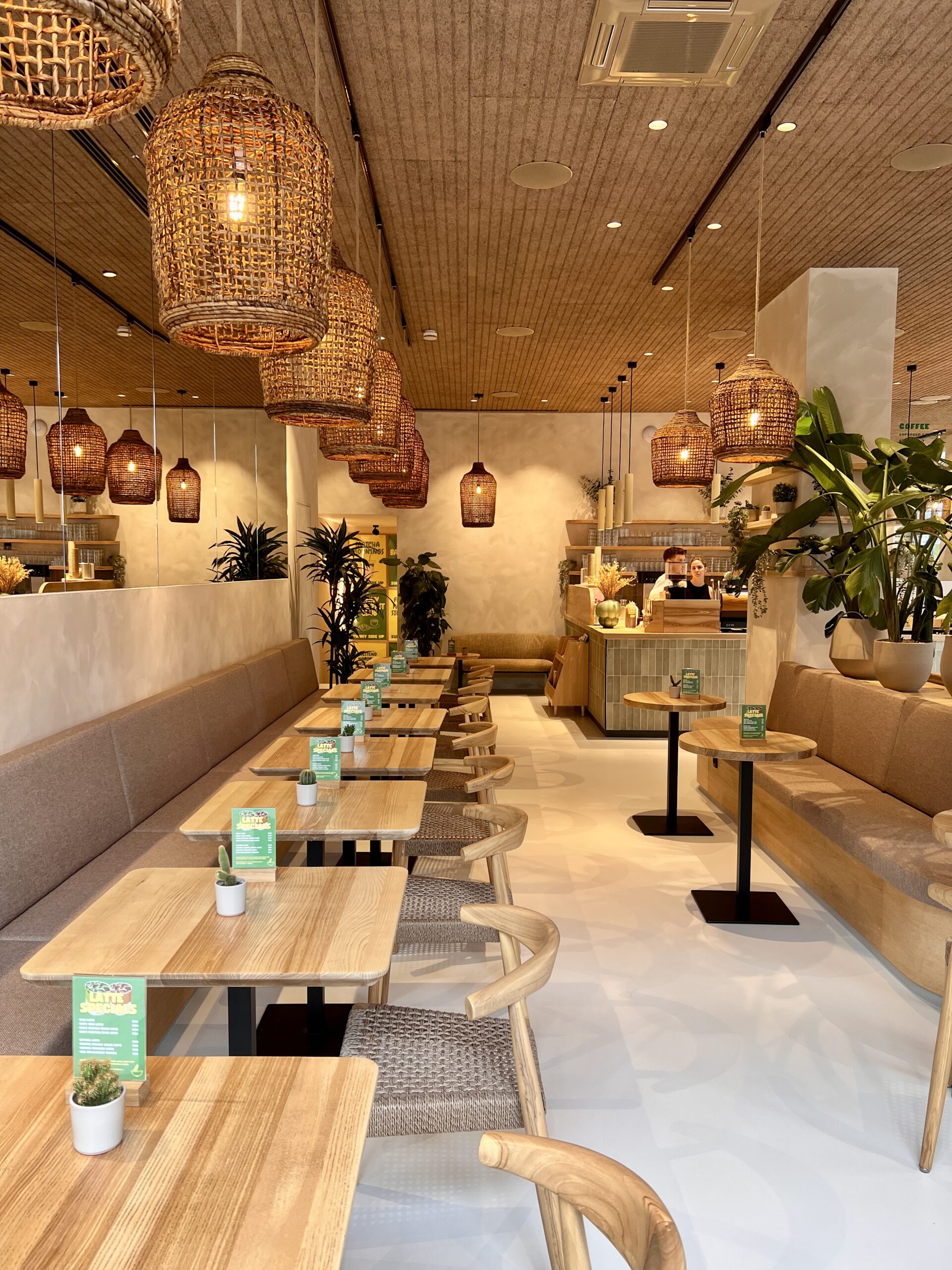 Modernes Café mit Holzstühlen, Tischen und stilvollen, geflochtenen Lampen; Pflanzen und ein Kaffeetresen im Hintergrund.