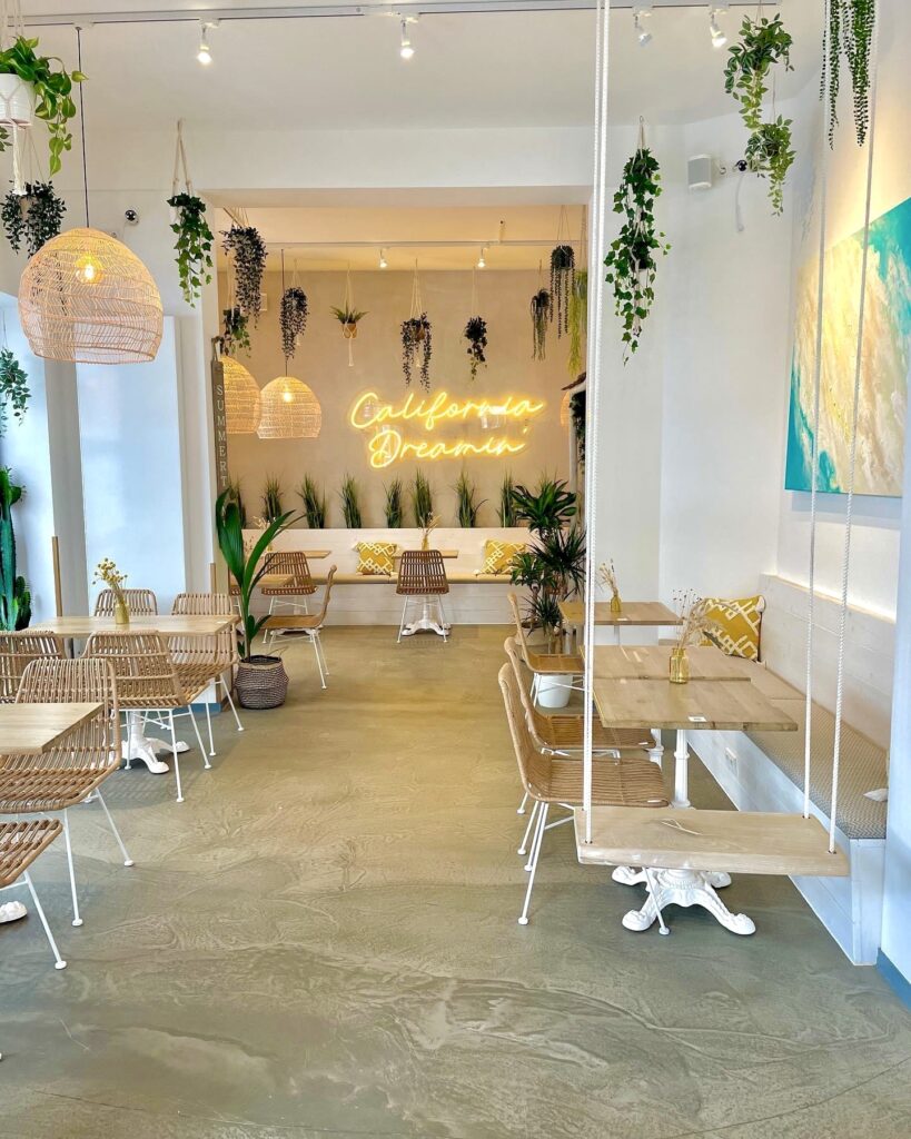 Helle Café-Atmosphäre mit Pflanzen, Rattanstühlen und einer neonbeleuchteten Wand mit dem Schriftzug „California Drea...