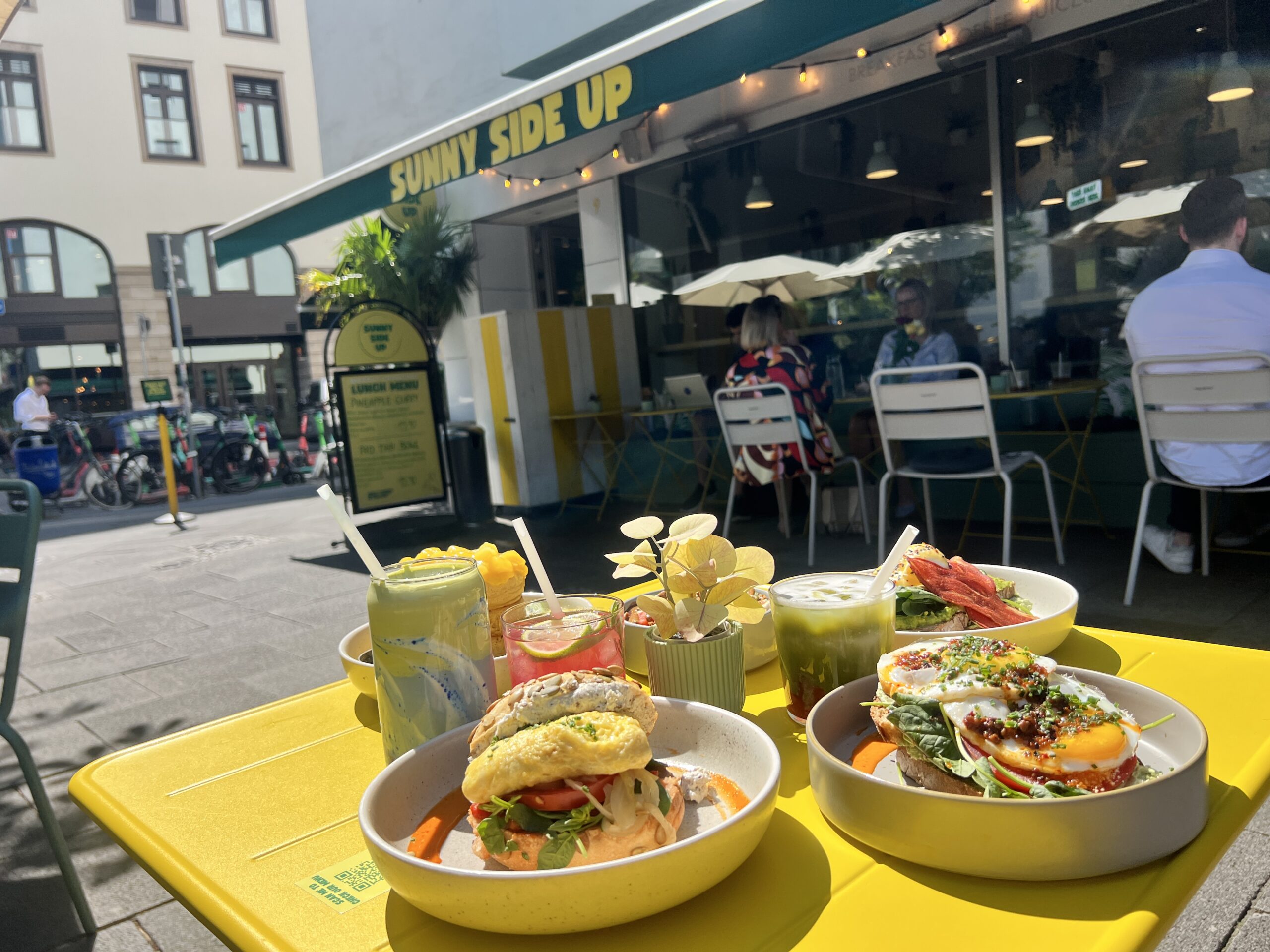 Bunt gedeckter Tisch mit verschiedenen Gerichten und Getränken vor einem Café namens "Sunny Side Up".