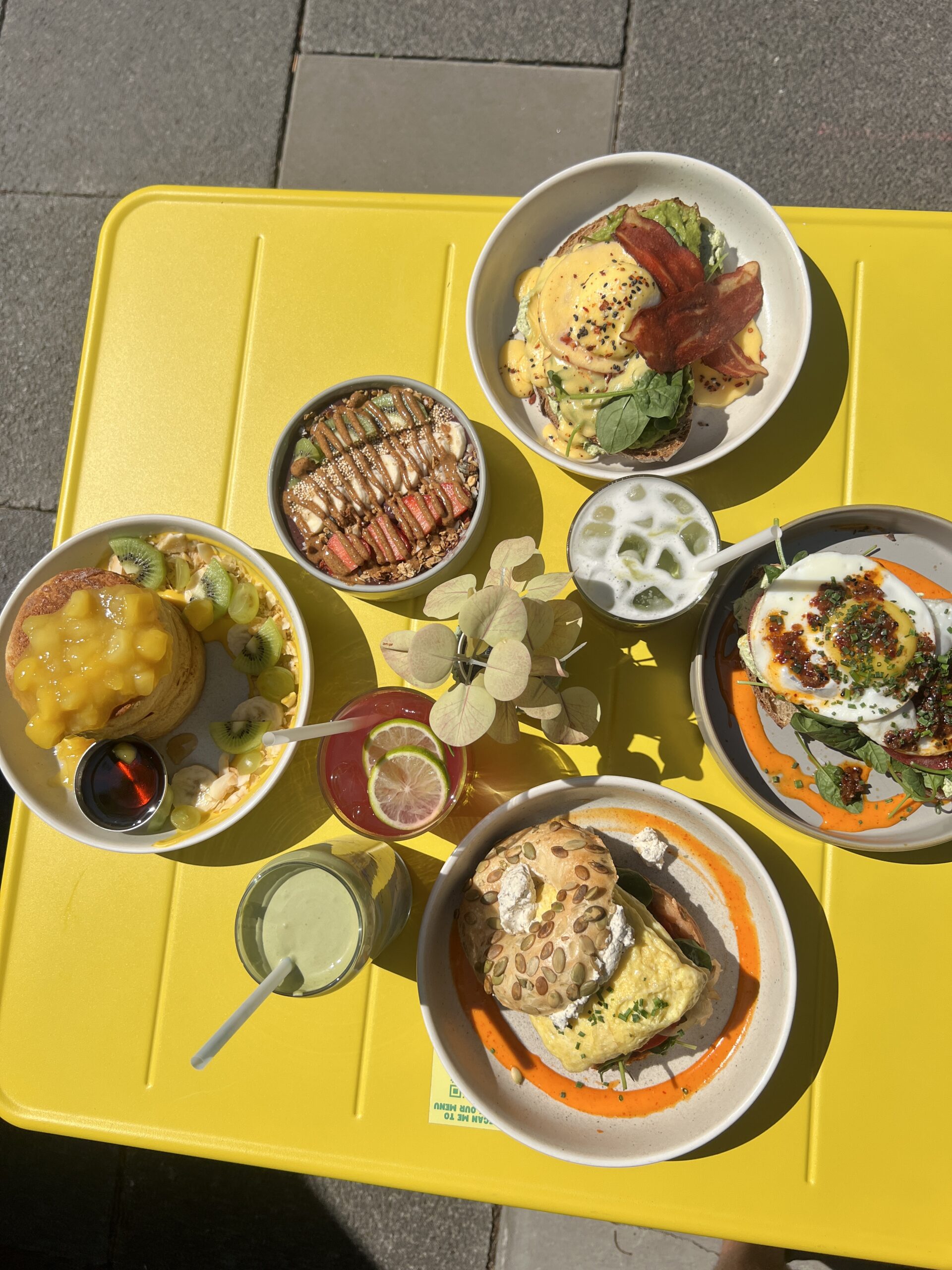 Bunt gedeckter Tisch mit verschiedenen Frühstücksgerichten, darunter Eier, Pancakes und Getränke auf einem gelben Tisch.