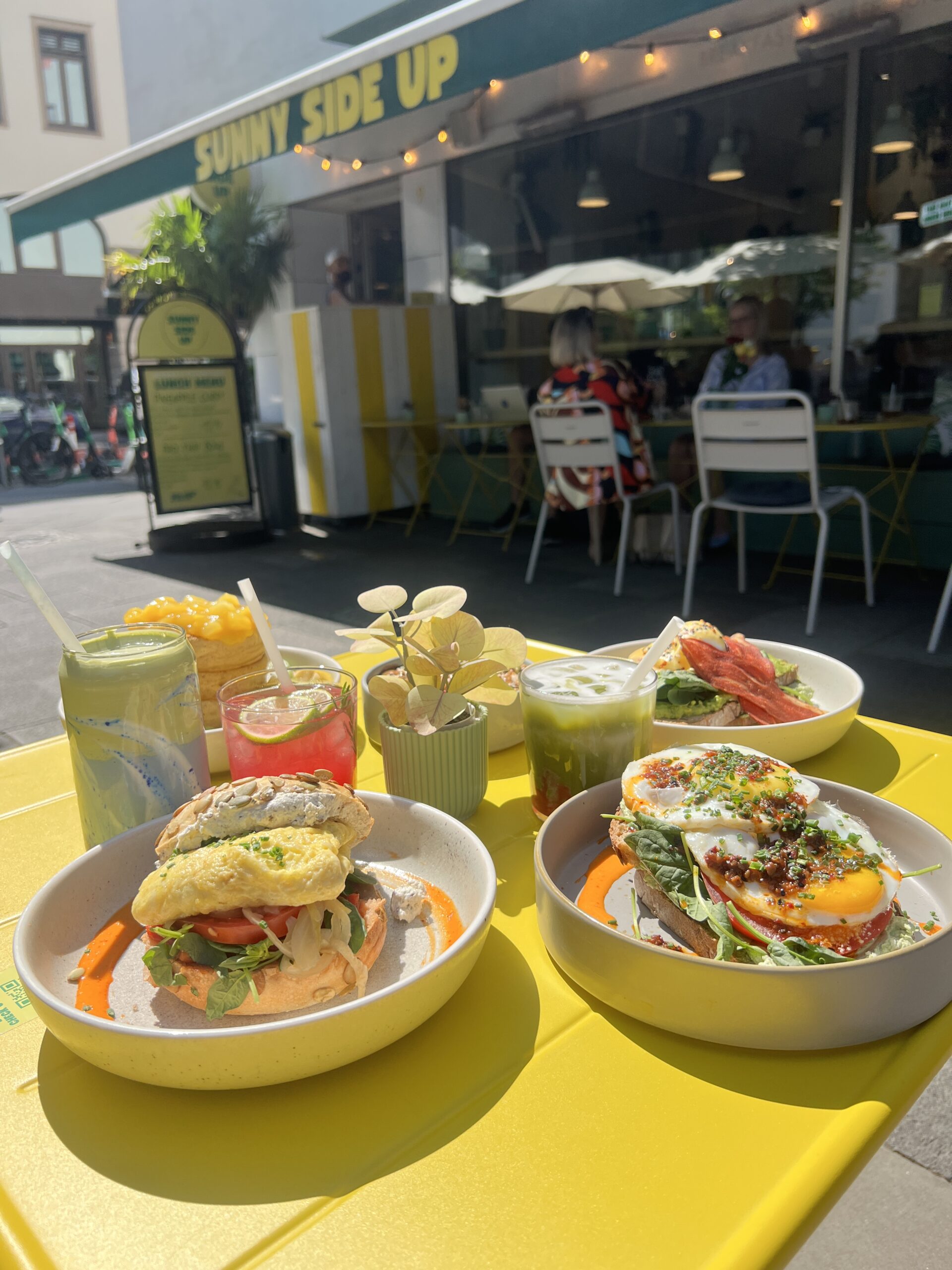 Tisch mit buntem Frühstück: Sandwich mit Omelett, verschiedene Getränke und einem Café im Hintergrund. Sonniges Wetter.