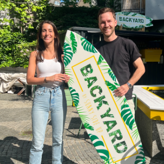 Sunny Side Up - Zwei Personen stehen lächelnd im Freien und halten ein Surfbrett mit der Beschriftung „BACKYARD“.