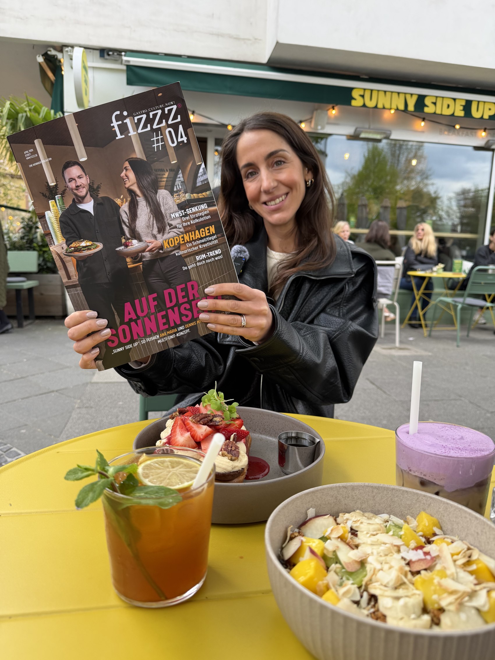 Ana Maria vor dem Sunny Side Up mit dem fizzz-Magazin und Fruehstueck auf dem Tisch